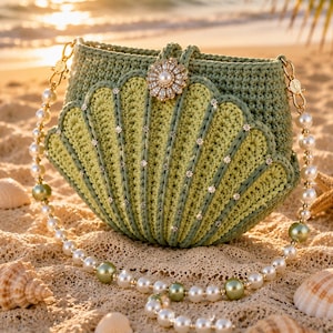 Op de afbeelding: Een saliegroene gehaakte handtas in de vorm van een schelp, voorzien van een centrale bloemvormige sluiting met een witte parel. De tas is versierd met kleine heldere kristallen langs de ribbels en heeft een schouderband van witte en groene parels.