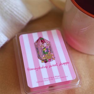 May include: A rectangular wax melt container with a pink and white striped label featuring a candy shop illustration and the text Honeydukes sweet shoppe, Second Star Waxes, Hand poured in the UK, and 100% coconut wax, positioned near a mug.