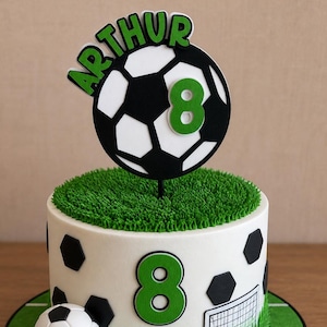 May include: A round cake decorated with a football theme, featuring a white base with black hexagon patterns, a green grass-textured top, a football topper with the name ARTHUR and number 8, a number 8 on the side, and a small football goal graphic.