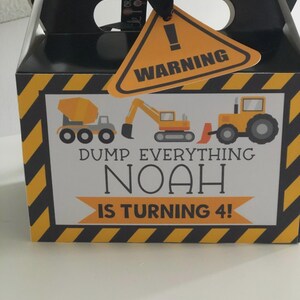 May include: A black cardboard favour box with a handle, featuring yellow and black diagonal caution stripes on the edges. The front displays construction vehicles and the text: DUMP EVERYTHING NOAH IS TURNING 4! A triangular orange warning tag is attached.