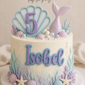 May include: A round cake with white frosting, decorated with a light blue shell topper featuring the number 5, a purple mermaid tail, and the name Isobel in purple script. The base features edible starfish, seashells, and coral in pastel tones.