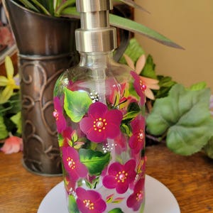 May include: A glass soap dispenser with a stainless steel pump. The dispenser is decorated with hand-painted pink flowers and green leaves.