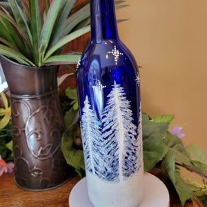 May include: A cobalt blue glass bottle with a white winter forest design. The bottle features a decorative stopper with a clear, faceted top. The bottle is decorated with white trees and star-like accents.