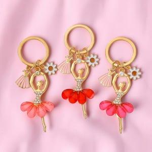 May include: Three gold-toned ballerina keychains with red and pink skirts. Each keychain features a ballerina charm with rhinestone accents, a small dress charm, and a white flower charm. The background is a light pink.