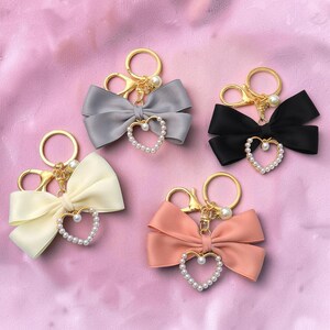 May include: Four keychains with satin bows in gray, black, cream, and peach. Each keychain has a gold-tone ring, a pearl accent, and a heart-shaped pendant outlined with small pearls. The background is a soft pink.