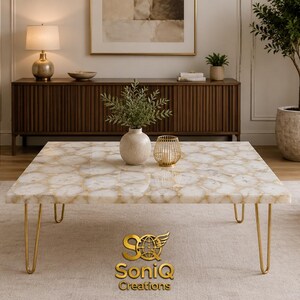 May include: A square coffee table with a white and gold patterned top and gold hairpin legs. The table is in a living room setting with a sofa, chairs, and a wooden cabinet. The logo "SoniQ Creations" is at the bottom.