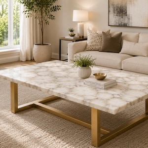 May include: A living room scene featuring a large, rectangular coffee table with a patterned, light-colored top and gold-colored metal legs. A beige sofa with decorative pillows sits behind the table. The room has a neutral color palette and a rug.