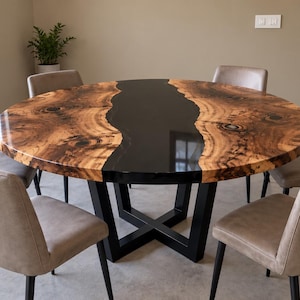 May include: A round dining table featuring a natural wood slab with a prominent live edge and a glossy black resin river inlay, supported by a black metal X-shaped base, surrounded by six beige upholstered chairs with black metal legs.