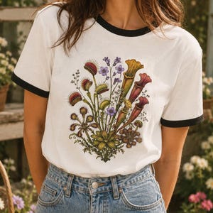 Könnte beinhalten: Ein weißes Ringer-T-Shirt mit schwarzem Besatz an Kragen und Ärmeln, das eine detaillierte botanische Illustration verschiedener fleischfressender Pflanzen zeigt, darunter Venusfliegenfallen, Kannenpflanzen und Sonnentau in Grün, Rot und Violett.