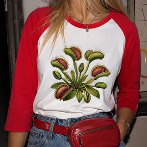 May include: A white baseball-style shirt with red three-quarter length sleeves and a red collar. The centre of the shirt features a detailed graphic illustration of a green and reddish-brown Venus flytrap plant with multiple open traps.