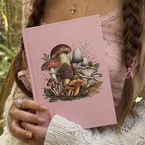 Puede incluir: Un libro de tapa dura de color rosa claro con una ilustración botánica detallada de varios hongos y follaje del bosque en la portada. El libro tiene un acabado mate suave y un pequeño marcapáginas de cinta rosa en el borde lateral.