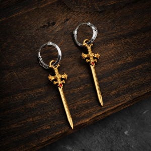 Può includere: Un paio di orecchini a cerchio con pendenti a forma di spada color oro. Ogni spada presenta un design elaborato dell'elsa con una piccola pietra preziosa rossa a taglio quadrato incastonata al centro. I cerchi sono color argento e circolari.