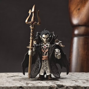 May include: A miniature vampire figurine, holding a gold trident. The figure is dressed in a black coat with ornate silver and gold detailing. The vampire has pale skin, black hair, and a stern expression. A small, red gem is visible on the chest.
