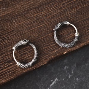 Peut inclure: Une paire de boucles d'oreilles créoles en argent en forme de serpent se mordant la queue. Le métal présente un motif d'écailles texturé et de petites pierres noires en guise d'yeux. Chaque boucle possède une petite bague avec du texte gravé.