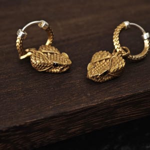 Puede incluir: Un par de pendientes de aro con un dije de cabeza de serpiente en tono dorado con texturas de escamas detalladas y pequeños ojos negros. Los aros tienen un patrón de escamas a juego y un mecanismo de bisagra plateado para el cierre.