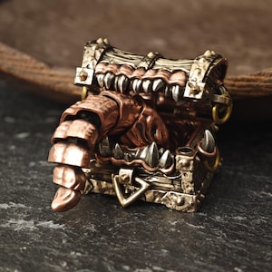 Puede incluir: Un cofre del tesoro detallado de estilo steampunk con un monstruo que emerge desde dentro. El cofre está hecho de bronce y cobre, con dientes afilados y un brazo segmentado. El diseño tiene un tema de fantasía.