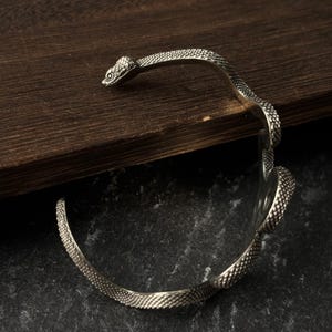 May include: A silver-toned metal cuff bracelet shaped like a coiled snake. The bracelet features a textured, scale-like pattern covering the entire surface, with a detailed snake head at one end and a curved tail section at the other end.
