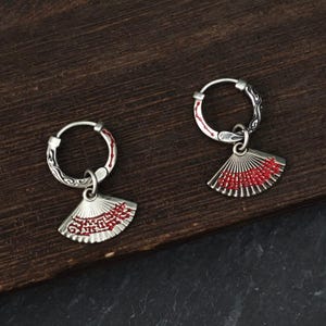 May include: A pair of silver hoop earrings featuring dangling fan-shaped charms. Each hoop has engraved scrollwork and a thin red line detail. The fan charms are silver with intricate red patterns across the surface, suspended by small silver rings.