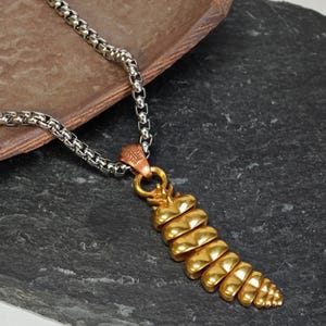 Puede incluir: Un collar con colgante dorado y cadena plateada. El colgante tiene un diseño segmentado en forma de cono. El collar se muestra sobre una superficie oscura y texturizada.