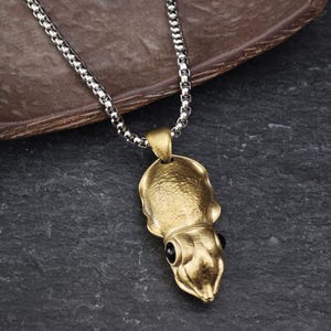 May include: A gold-colored squid pendant necklace with a silver chain. The squid pendant has black eyes and is detailed with textured surfaces. The necklace is displayed on a dark, textured surface.