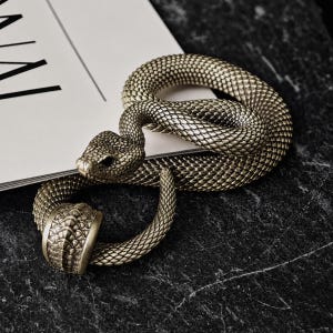 Puede incluir: Una figura de serpiente enroscada hecha de un material metálico en tonos bronce con un patrón de escamas detallado y texturizado en todo el cuerpo. La serpiente está colocada en un bucle circular, con un segmento de banda ancho y liso cerca de la cola.