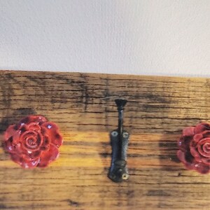 May include: A rustic wooden board with various hardware elements. It features two red rose-shaped knobs, two metal knobs, and a black metal latch. The wood has a natural, weathered appearance, with visible grain and knots.