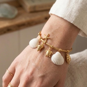 May include: Gold-toned bracelet featuring seashell charms, a starfish, and a small turtle pendant. The bracelet includes white seashell charms and is worn on a wrist with multiple gold rings.