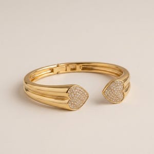 May include: A gold-colored open cuff bracelet with heart-shaped ends. The heart-shaped ends are encrusted with small, sparkling stones. The bracelet has a smooth, polished finish and a hinged clasp.