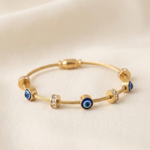 Pode incluir: Pulseira dourada com amuletos de olho grego azul e detalhes brilhantes. A pulseira tem um design delicado, semelhante a um fio, com um fecho magnético. O diâmetro da pulseira é de aproximadamente 6,35 cm.