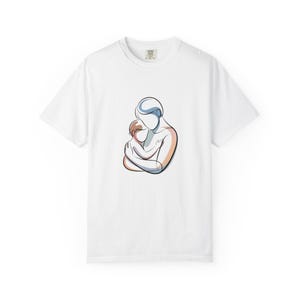 Peut inclure: T-shirt blanc avec une illustration minimaliste d'un parent embrassant un enfant. L'œuvre utilise des couleurs douces, notamment des nuances de marron, d'orange et de bleu. Le dessin est centré sur le t-shirt.