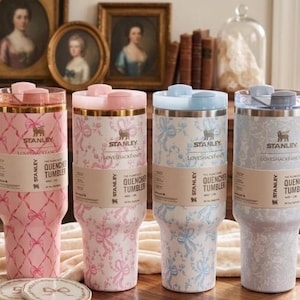 K&ouml;nnte beinhalten: Vier Stanley Quencher-Becher in Rosa, Hellblau und Grau, jeweils mit unterschiedlichen Blumen- und Schleifenmustern. Die Becher haben passende Deckel und der Text "Stanley Loveshackfancy Quencher Tumbler" ist sichtbar.