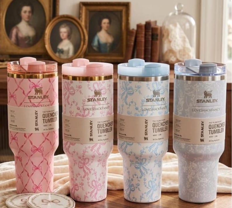 K&ouml;nnte beinhalten: Vier Stanley Quencher-Becher in Rosa, Hellblau und Grau, jeweils mit unterschiedlichen Blumen- und Schleifenmustern. Die Becher haben passende Deckel und der Text "Stanley Loveshackfancy Quencher Tumbler" ist sichtbar.