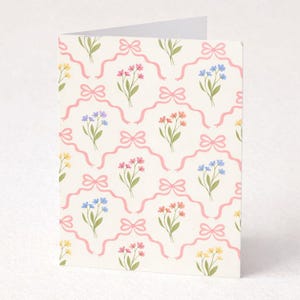 May include: A folded greeting card with a repeating pattern of pink bows and colorful floral bouquets. The flowers are in shades of yellow, pink, blue, and orange, set against a cream background. The card is blank inside.