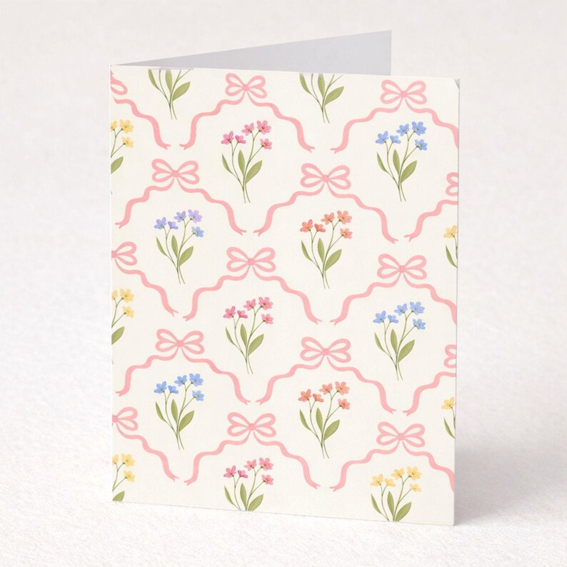 May include: A folded greeting card with a repeating pattern of pink bows and colorful floral bouquets. The flowers are in shades of yellow, pink, blue, and orange, set against a cream background. The card is blank inside.