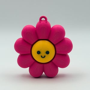 May include: A vibrant pink flower charm with a yellow centre and a smiling face. The charm has a loop for hanging. The flower has eight petals and is likely made of plastic. A cheerful design.