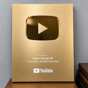 Puede incluir: Una placa rectangular de color dorado del premio YouTube Creator Award con un icono de botón de reproducción central. El texto dice: Presented to Urban Therapy BE For passing 1,000,000 subscribers, con el logo de YouTube impreso en la parte inferior.