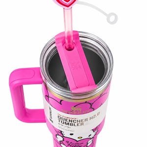 May include: A pink Stanley Quencher H2.0 40 oz tumbler featuring a Hello Kitty graphic, a pink handle, and a clear lid with a pink straw topped by a heart-shaped accessory. The label displays the text The Flowstate Quencher H2.0 40 oz 1.18 L.
