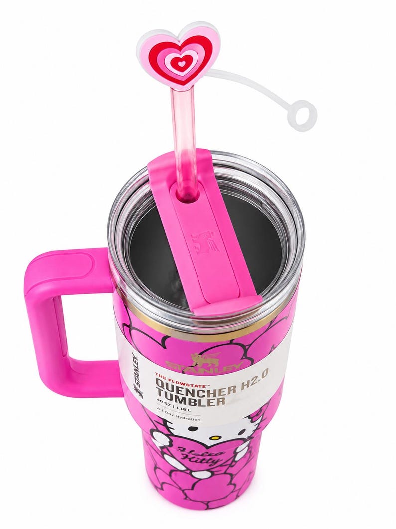 May include: A pink Stanley Quencher H2.0 40 oz tumbler featuring a Hello Kitty graphic, a pink handle, and a clear lid with a pink straw topped by a heart-shaped accessory. The label displays the text The Flowstate Quencher H2.0 40 oz 1.18 L.