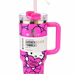 May include: A bright pink 40 oz Stanley Quencher H2.0 tumbler featuring a Hello Kitty graphic, black heart outlines, and a gold rim. The cup includes a pink handle, a clear lid, and a straw topped with a pink and white heart-shaped accessory.