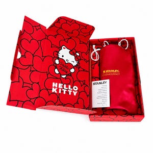 May include: A red rectangular gift box featuring a Hello Kitty graphic holding a heart and a heart pattern. Inside, a red satin drawstring pouch with the gold text Stanley LovesHackFancy and a white Stanley instruction card are visible.