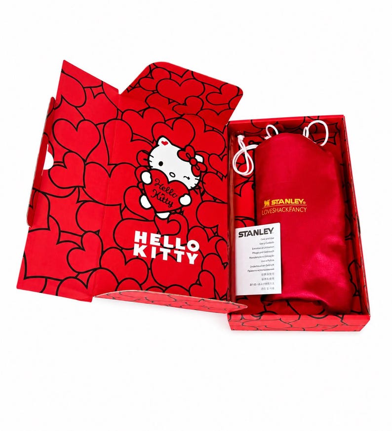 May include: A red rectangular gift box featuring a Hello Kitty graphic holding a heart and a heart pattern. Inside, a red satin drawstring pouch with the gold text Stanley LovesHackFancy and a white Stanley instruction card are visible.