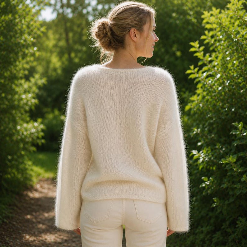 May include: A long-sleeved, cream-colored knit sweater with a soft, fuzzy texture and a wide neckline. The sweater features a simple, solid-colored design with ribbed detailing along the collar and cuffs, paired with matching cream-colored trousers.