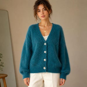 Op de afbeelding: Een blauwgroen vest met V-hals en een pluizige, getextureerde gebreide afwerking. Het kledingstuk heeft lange mouwen met geribbelde boorden, een geribbelde zoom en een knoopsluiting aan de voorzijde met vier lichtgekleurde knopen.
