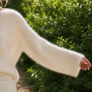 May include: A long-sleeved, off-white sweater with a fuzzy, brushed texture. The garment features a relaxed fit with dropped shoulders and wide sleeves, crafted from a soft, knit material that creates a light, airy appearance across the fabric.