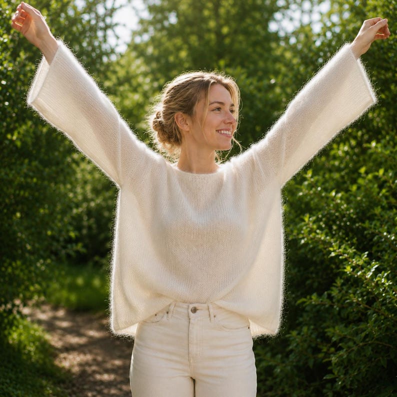 May include: A cream-colored long-sleeve sweater with a fuzzy, soft-textured knit, a wide round neckline, and loose, flared sleeves. The sweater is paired with matching cream-colored pants, creating a monochromatic look with a relaxed fit.