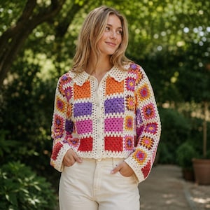 May include: A long-sleeved crochet cardigan featuring a colorful granny square pattern in shades of orange, purple, pink, and red, accented with cream-colored borders. The garment includes a scalloped collar and a front button closure.