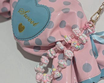 Può includere: Una borsa a mano rosa e blu a pois con una toppa a forma di cuore che dice "Sweet" e un ciondolo fatto di perline color pastello.