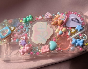 May include: A clear phone case decorated with pastel-colored charms, including hearts, stars, candies, and a bear. The case is filled with glitter and rhinestones.
