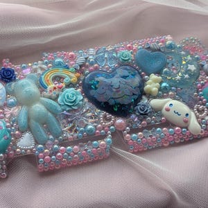 May include: A blue and pink phone case decorated with a variety of charms, including a rainbow, a heart, a bear, and a cartoon character. The case is covered in glitter and rhinestones.