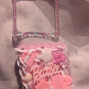 May include: A pink and white clear plastic case with a pink crown on top. The case is filled with pink glitter and has a variety of pink and white charms, including a star, a heart, a rose, a purse, a comb, and the word "Barbie".